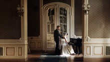 Elegant Stylish Handsome Groom Looking At His Gorgeous Bride Playing The Piano. Reflection In The Mirror. Unusual Luxury Wedding Couple In Retro Style