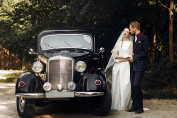 Wall Mural - luxury elegant wedding couple holding hands on background of stylish black car .  gorgeous bride and handsome groom in retro style. romantic moment.