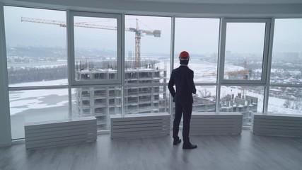 businessman in the white office looking on the street with construction site wearing in elegant suit drinking coffee oversees how workers working. rear back view man with orange hard hat in modern