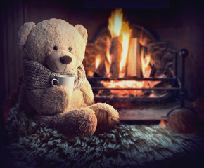 a teddy bear is sitting by the fireplace with a cup