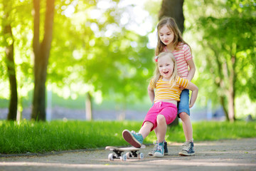 Wall Mural - Two pretty little girls learning to skateboard on beautiful summer day in a park