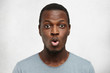 Close up shot of funny young African American man dressed casually pouting lips and raising brows, expressing shock or surprise, posing against white wall background with copy space for your text