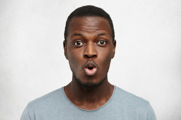 Wall Mural - Close up shot of funny young African American man dressed casually pouting lips and raising brows, expressing shock or surprise, posing against white wall background with copy space for your text