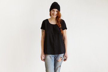 Wall Mural - Indoor shot of beautiful happy teenager girl with tattoos wearing her ginger hair in messy braid, standing isolated against blank copy space wall, dressed in ragged jeans, t-shirt and trendy hat