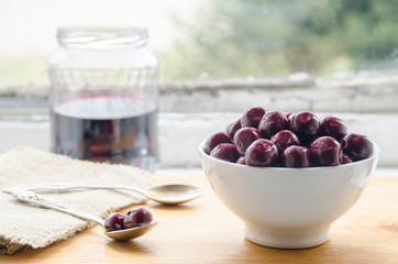 Cherries from compote in a white ceramic bowl, two silver spoons and a jar with cherry juice in the background