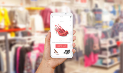 Wall Mural - Online shopping with mobile phone. PHone in woman hand. Shop market online app on screen. Clothes and footwear shop in background.