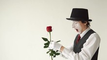 Man Mime With A Red Rose On A White Background. MIM Fingers Pricked On The Thorn Of A Rose