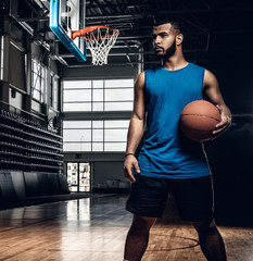 Wall Mural - Black basketball player holds a ball over a hoop in a basketball hall.