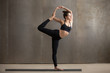 © fizkes - Young attractive smiling yogi woman practicing yoga, standing in Natarajasana exercise, Lord of the Dance pose, working out wearing black sportswear, urban style grey studio background, full length