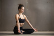 © fizkes - Young attractive sporty yogi woman practicing yoga, sitting in Sukhasana exercise, Easy Seat pose, working out wearing black sportswear, cool urban style grey studio copy space background, full length