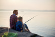 © pressmaster - Back view portrait of adult man and teenage boy sitting together on rocks fishing with rods in calm waters of blue lake at dusk, both wearing checkered shirts