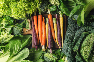 Variety of raw green vegetables salads, lettuce, bok choy, corn, broccoli, savoy cabbage, colorful young carrots on wooden chopping board. Food background. Top view, space for text