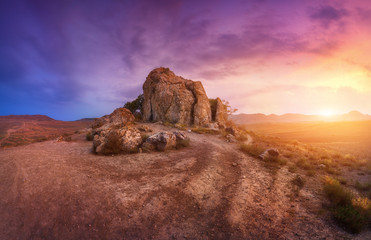 Rocks against amazing cloudy sky in desert at sunset. Blue, purple and red clouds. Panoramic. Colorful landscape with trail, stones, grass and yellow sunlight. Nature and travel background. Adventure