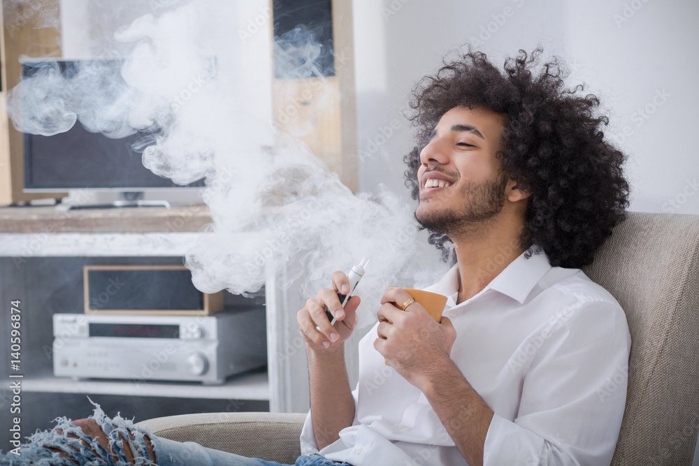 Weekends concept. Hipster man smoking his favorite electronic cigar and  drinking coffee or tea while sitting in arm chair at home alone. Stock  Photo