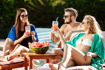 Wall Mural - Friends smiling, eating watermelon, drinking cocktails, relaxing near swimming pool.