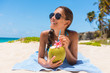 Sunglasses beach woman drinking coconut water having fun on summer vacation. Tropical travel holidays. Bikini casual girl relaxing lying down with natural healthy drink.