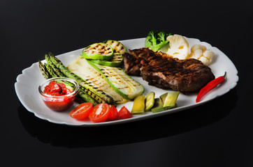 Wall Mural - Grilled beef grilled steak with asparagus, zucchini, lobi, broccoli, tomato, chili and red sauce on a white plate, on a black glossy background