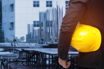 Wall Mural - double exposure of businessman hold in hand yellow safety helmet  industrial concept on blurred construction site workers background, color tone effect.