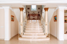 Luxurious Staircase With Marble Steps And Decorative And  Ornamental Iron Railings. On The Top Are Decorating Furniture In Neoclassical Style, A Table With Candles. Corinthian Column Style.