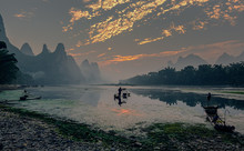 Fisherman Stands On Traditional Bamboo Boats At Sunrise (boat With A Red Sail In The Background) - The Li River, Xingping, China