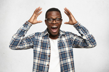 Wall Mural - I can't believe this. Amazed young African American hipster wearing trendy glasses and checkered shirt over white t-shirt holding hands in surprised gesture, keeping mouth wide open, looking shocked