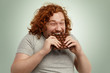Plump funny redhead young Caucasian male with curly hair biting large of chocolate with pleased joyful look, keeping his eyes closed, enjoying its sweet taste after week of strict vegetarian diet