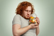 Obese chubby European man with ginger curly hair holding jar of sweets tight, having greedy look, frowning and grimacing, posing indoors at studio wall. Obesity, gluttony and unhealthy lifestyle