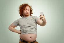 Funny Overweight Plump Man With Duck Lips Wearing Undersize T-shirt With Belly Hanging Out Of Pants, Keeping Hand On Waist, Posing For Selfie, Holding Cell Phone, Trying To Seem Attractive And Sexy