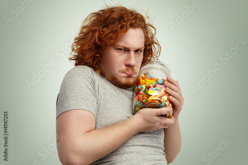 Fotografía Obese chubby European man with ginger curly hair holding jar of sweets tight, having greedy look, frowning and grimacing, posing indoors at studio wall