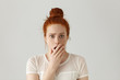 Portrait of surprised amazed attractive young redhead female wearing white top having astonished face expression, covering her open mouth with hand, looking at camera in shock and full disbelief