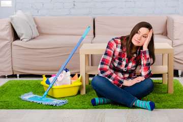 exhausted woman dressed in casual clothes sitting on the floor, during housework. Beside her cleaning supplies, mop for cleaning the house.