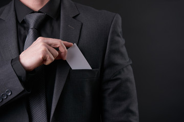 Young Successful Businessman in black suit and gray necktie put or take out visit card in pocket on isolated black background