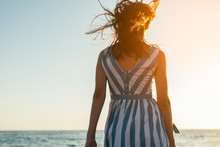 Back View Silhouette Of Brunette Beautiful Woman Walking Along Beach And Sea Sunset Background. Cover Idea. Female In Dress Walking On Ocean, Wind Blowing Hair. Loneliness And Sadness Concept.