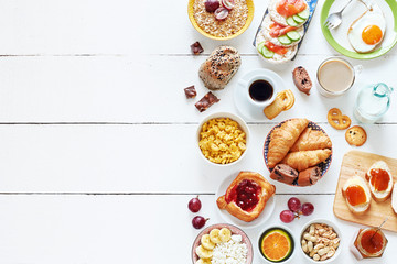 Breakfast menu food frame with a copy space. Eggs, croissants, oatmeal with banana, cereal, danish pastry, crisps with salmon, apricot jam, fresh bread, chocolate, coffee and milk on a white table.