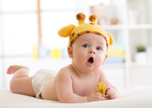 Funny Baby Boy In Giraffe Hat Lying On His Belly In Nursery. Little Kid Lies On Bed With Opened Mouth And Holds Soother In Hands