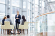 © pressmaster - Group of four business people sitting in armchairs during meeting, one woman giving presentation in glass hall of modern office building