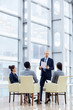 © pressmaster - Group of  business people sitting in circle during meeting, man standing up giving presentation in glass hall of modern office building