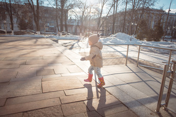 little girl in a beige jacket, blue jeans and pink rubber boots running in a park in the early spring on a sunny day, melting snow and blue sky, building