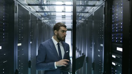 Wall Mural - IT Engineer in Data Center Stands Before Server Rack Cabinet Working on His Laptop. Runs Diagnostics or Maintenance Job. Shot on RED EPIC-W 8K Helium Cinema Camera.