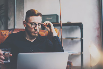 Pensive bearded businessman in eyeglasses working at modern loft.Man sitting in vintage chair,holding in hands glass of water,using contemporary notebook.Blurred background,flares.Horizontal