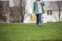 Low Section Of Girl Carrying Watering Can And Umbrella While Walking On Grassy Field