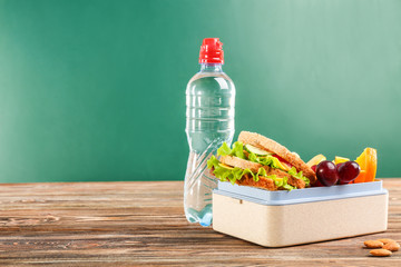 Appetizing food in lunch box and bottle of water on wooden table against chalkboard background