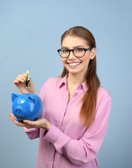 Canvas Print - Beautiful young woman putting money into piggy bank, on color background