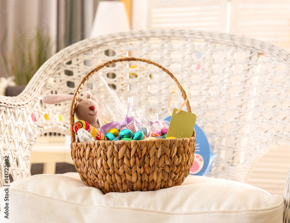 Beautiful Easter basket with traditional decorations and sweets on wicker chair in light room