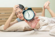 © praisaeng - Sleepy young woman in bed with eyes closed extending hand to alarm clock at home