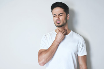 Young man having sore throat and touching his neck, wearing a loose white t-shirt against light grey background. Hard to swallow