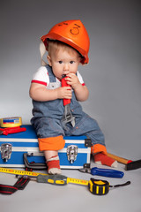 child in an orange construction helmet sits on a toolbox and gnaws the adjustable wrench. Around on the floor are scattered tools: tape measure, hammer, screwdriver