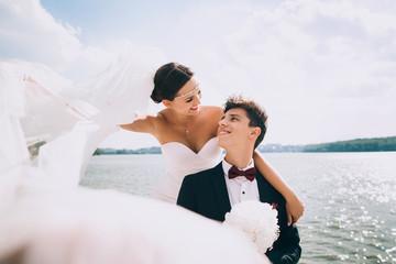 Elegant smiling young bride and groom walking on the beach, kissing and having fun, wedding ceremony near the lake