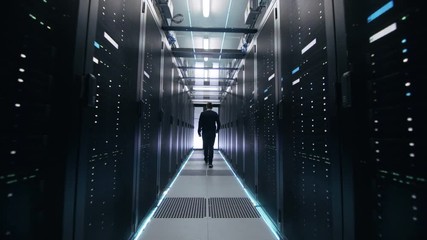 Wall Mural - Following Shot of IT Engineer Walking Through Data Center Corridor with Rows of Rack Servers. Opens Laptop. Shot on RED EPIC-W 8K Helium Cinema Camera.