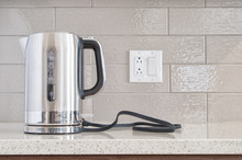 Modern Electric Stainless Steel Kettle Sitting On Its Base On A Granite Counter Top Against A Ceramic Background